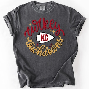May include: Dark grey t-shirt featuring the words "turkey touchdowns" in red and gold script. A white arrowhead with "KC" in red is in the center of the design. The shirt is folded.