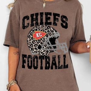 Könnte beinhalten: Braunes T-Shirt mit dem Schriftzug "CHIEFS FOOTBALL" in schwarzen Blockbuchstaben. Ein Football-Helm mit Leopardenmuster und den Buchstaben "KC" in einem roten Pfeil befindet sich in der Mitte. Das Shirt hat eine lockere Passform.