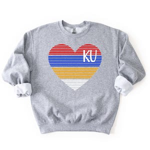 May include: A heather gray sweatshirt featuring a heart design with red, blue, yellow, and white stripes, and the letters "KU" in white. The sweatshirt has long sleeves with rolled-up cuffs.