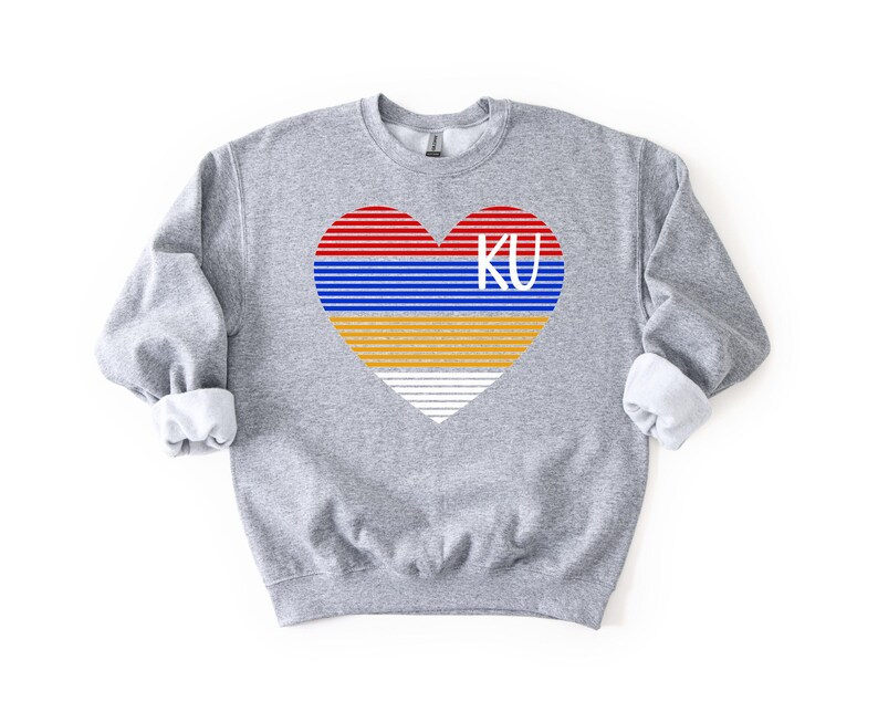 May include: A heather gray sweatshirt featuring a heart design with red, blue, yellow, and white stripes, and the letters "KU" in white. The sweatshirt has long sleeves with rolled-up cuffs.