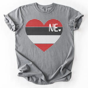 May include: A gray t-shirt featuring a heart design with red, black, white, and gray horizontal stripes. The letters "NE" are in white within the heart, with a small heart symbol below.