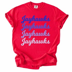 May include: A red t-shirt with the word "Jayhawks" printed in blue and white script four times down the front. The shirt is made of a soft material and has rolled-up sleeves.