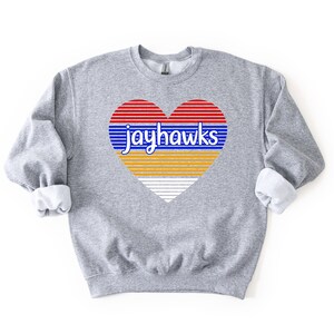 May include: A gray sweatshirt with a heart-shaped graphic. The heart design features red and blue horizontal stripes, with the word "jayhawks" in blue. The bottom half of the heart is yellow and white striped. The sweatshirt has a crew neck and long sleeves.