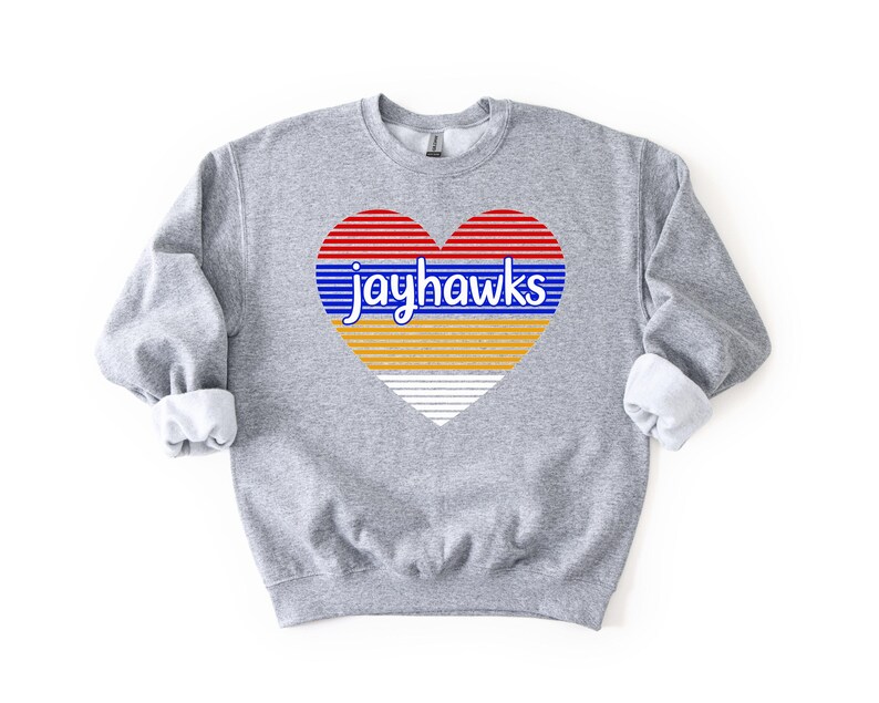 May include: A gray sweatshirt with a heart-shaped graphic. The heart design features red and blue horizontal stripes, with the word "jayhawks" in blue. The bottom half of the heart is yellow and white striped. The sweatshirt has a crew neck and long sleeves.