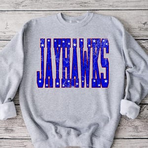 May include: A grey sweatshirt featuring the word "JAYHAWKS" in large, distressed blue letters with red accents and white stars. The sweatshirt has a crew neck and long sleeves with rolled-up cuffs.