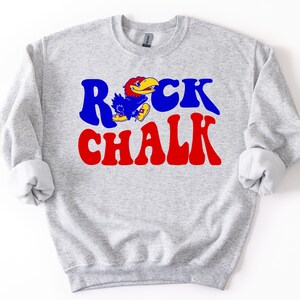 Puede incluir: Una sudadera gris claro con el texto "ROCK CHALK" en azul y rojo, con un gráfico de la mascota Jayhawk de dibujos animados. La sudadera tiene mangas largas con puños enrollados.