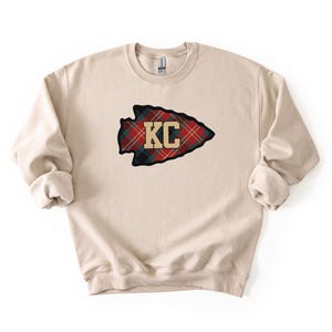 May include: A beige crewneck sweatshirt featuring a plaid arrowhead design with the letters "KC" in gold. The arrowhead is outlined in black and has a red, green, and black plaid pattern.