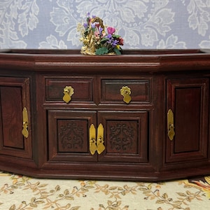 May include: A dark wood cabinet with two doors and two drawers. The cabinet has ornate carvings and brass hardware. A small floral arrangement sits on top of the cabinet.