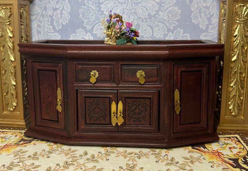 May include: A dark wood cabinet with two doors and two drawers. The cabinet has ornate carvings and brass hardware. A small floral arrangement sits on top of the cabinet.
