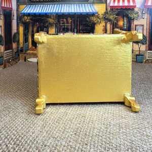 May include: A gold-painted wooden box with decorative feet. The box is sitting on a hessian surface.