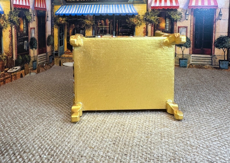 May include: A gold-painted wooden box with decorative feet. The box is sitting on a hessian surface.