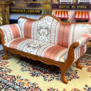 Puede incluir: Sofá en miniatura de estilo antiguo con estructura de madera y tapicería estampada. El sofá presenta un marco marrón claro y cojines con un diseño floral blanco, melocotón y verde. Es un mueble de casa de muñecas.