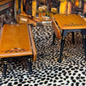 May include: Two miniature wooden tables with drop leaves. The tables have a warm wood finish with black legs and gold accents. Floral details are painted on the tabletop. One table is folded, and the other is open. The tables are on a leopard print surface.