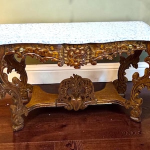 May include: Ornate, antique wooden bench with a white speckled marble top. The bench features intricately carved floral and scrollwork designs, with curved legs and a lower shelf. The wood has a warm, golden-brown finish.