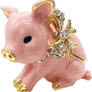 Puede incluir: Una figurita de cerdo rosa con un lazo dorado adornado con diamantes de imitación. El cerdo está sentado con las patas metidas debajo. La figurita es una caja de joyería.