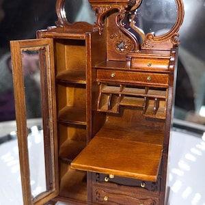 May include: A miniature wooden cabinet with a mirrored top and ornate carvings. The cabinet has a glass-doored section, shelves, drawers, and a drop-down desk. The wood has a warm, brown finish, and the cabinet is set on small, decorative feet.