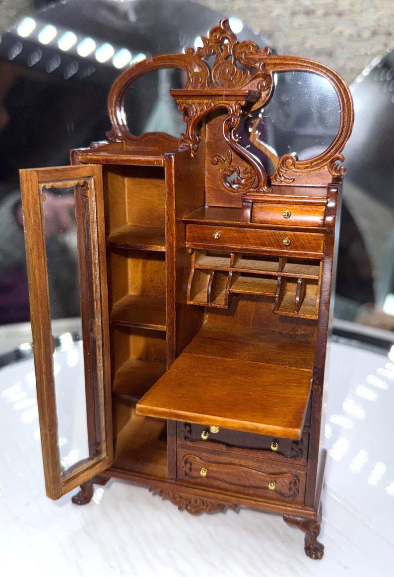May include: A miniature wooden cabinet with a mirrored top and ornate carvings. The cabinet has a glass-doored section, shelves, drawers, and a drop-down desk. The wood has a warm, brown finish, and the cabinet is set on small, decorative feet.