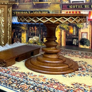 May include: A wooden table with a decorative gold trim and a carved base. The table is set on a patterned rug in front of a painted backdrop of a Parisian street scene.