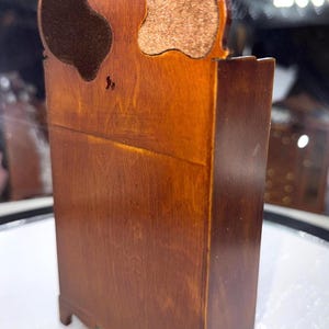 May include: A miniature wooden cabinet with a dark brown finish. The cabinet has a decorative top with two curved cutouts, one with a dark brown material and the other with a tan material. The cabinet is approximately 6 inches tall.