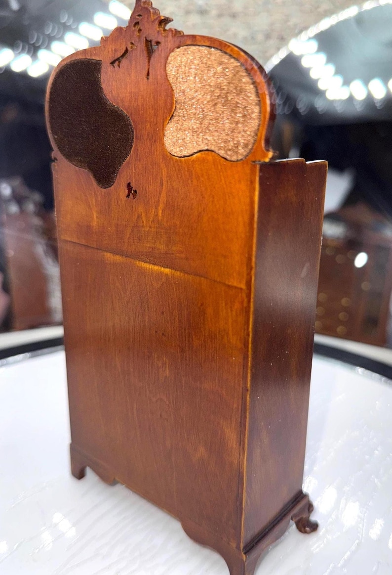 May include: A miniature wooden cabinet with a dark brown finish. The cabinet has a decorative top with two curved cutouts, one with a dark brown material and the other with a tan material. The cabinet is approximately 6 inches tall.