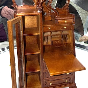 May include: A miniature wooden vanity with a mirror, drawers, and shelves. The vanity is made of dark brown wood with ornate carvings and gold-colored hardware. The cabinet door is open, revealing shelves.