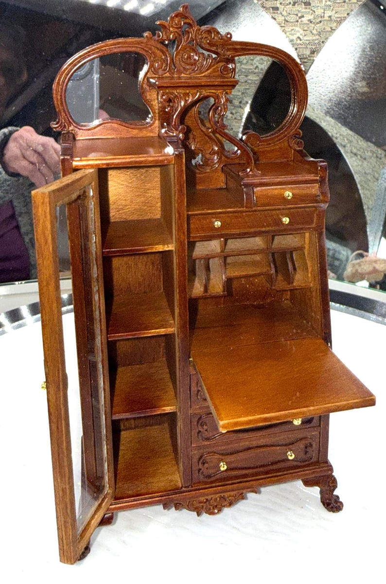 May include: A miniature wooden vanity with a mirror, drawers, and shelves. The vanity is made of dark brown wood with ornate carvings and gold-colored hardware. The cabinet door is open, revealing shelves.