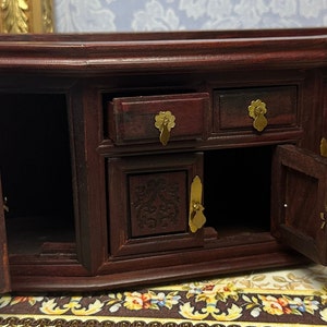May include: A dark wood miniature cabinet with two doors and three drawers. The doors are open, revealing the empty interior. The cabinet has brass handles and decorative carvings.