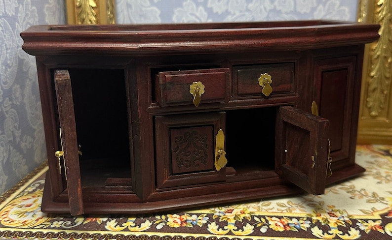 May include: A dark wood miniature cabinet with two doors and three drawers. The doors are open, revealing the empty interior. The cabinet has brass handles and decorative carvings.