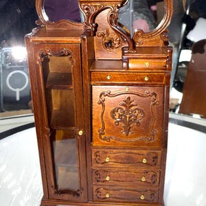 May include: Miniature wooden armoire with a mirrored vanity. The armoire features a glass-fronted cabinet, drawers, and ornate carvings. The wood has a rich, dark brown finish, and the hardware is gold-toned. The overall design is reminiscent of antique furniture.