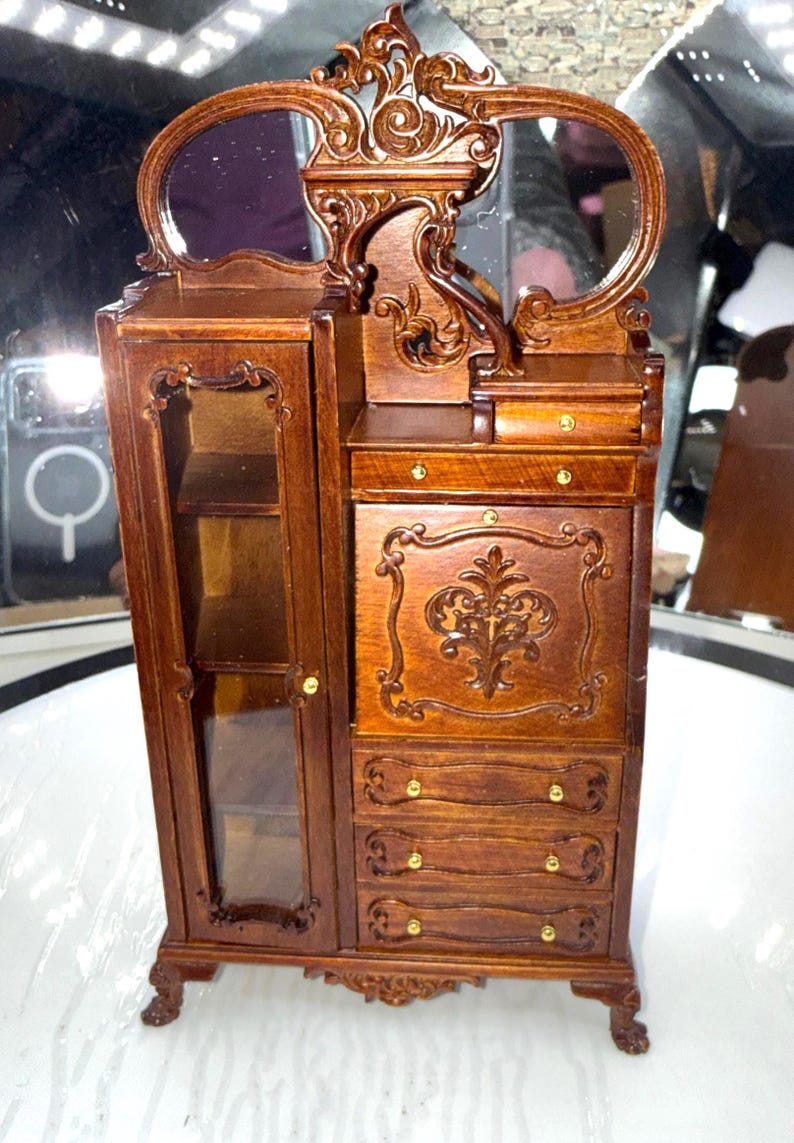 May include: Miniature wooden armoire with a mirrored vanity. The armoire features a glass-fronted cabinet, drawers, and ornate carvings. The wood has a rich, dark brown finish, and the hardware is gold-toned. The overall design is reminiscent of antique furniture.