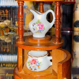 May include: A miniature wooden tea cart with a small white porcelain creamer and sugar bowl. The creamer and sugar bowl are decorated with pink and blue floral patterns. The cart has a dark brown finish.