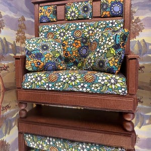 May include: Miniature wooden sofa with floral patterned cushions in shades of blue, orange, and green. The sofa has a dark brown wooden frame and a matching base. The cushions feature a repeating floral design.