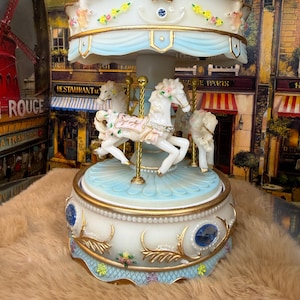 May include: A decorative carousel music box with white horses, gold accents, and a light blue canopy. The carousel features floral and jewel embellishments, creating a whimsical and elegant design. The base is adorned with intricate details, adding to its charm.