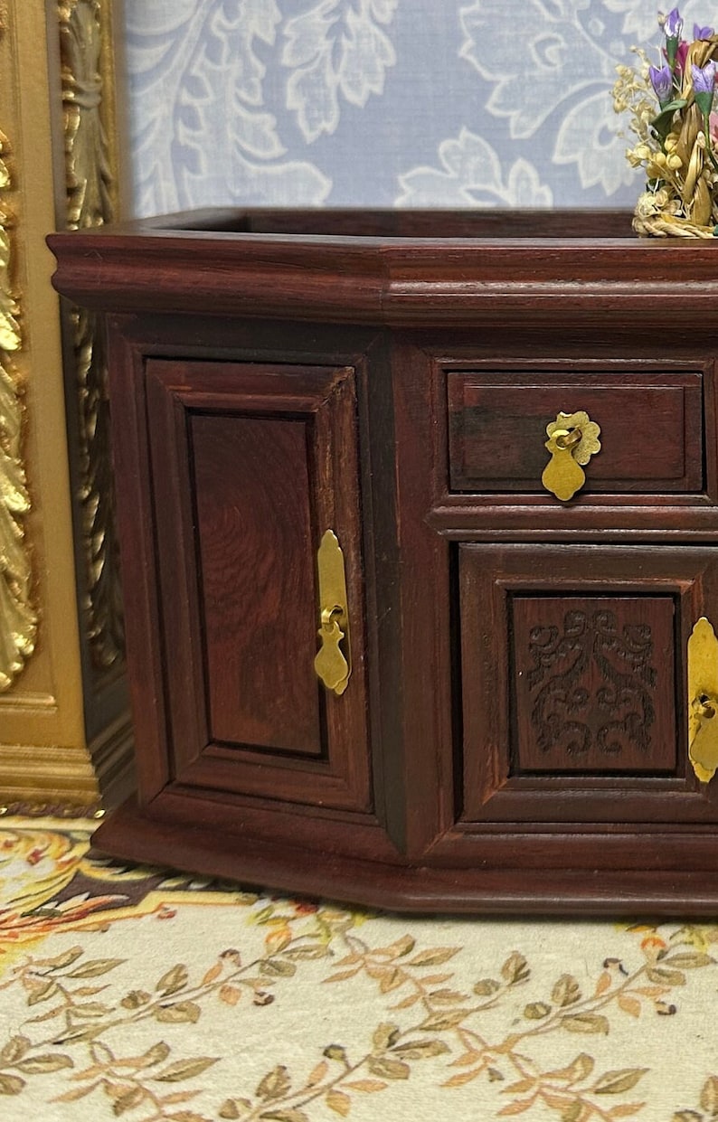 May include: A dark wood jewelry box with two doors and three drawers. The box has gold-colored handles and a carved design on the front.