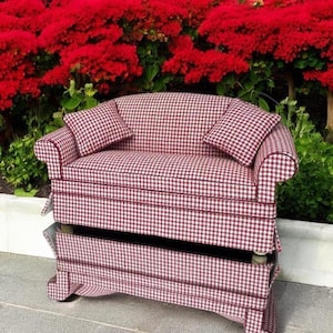May include: A miniature sofa upholstered in a red and white gingham fabric, with two matching cushions. The sofa is positioned in front of a backdrop of red flowers and green plants. The sofa has a classic design.