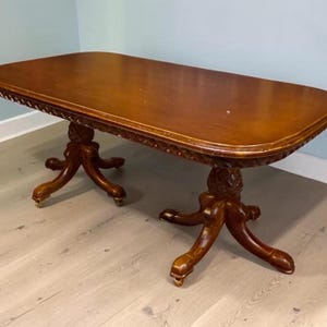 Op de afbeelding: Een rechthoekige houten salontafel met een glad, gepolijst bruin oppervlak en afgeronde hoeken. De tafel heeft een sierlijk gesneden rand en rust op twee voetstukken, elk met vier gebogen poten die eindigen in kleine metalen wieltjes.