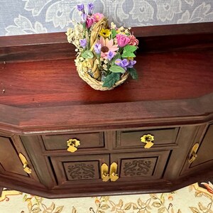 May include: A dark wood cabinet with two doors and two drawers. The cabinet has a carved design on the front and gold-colored handles. A basket of flowers sits on top of the cabinet.