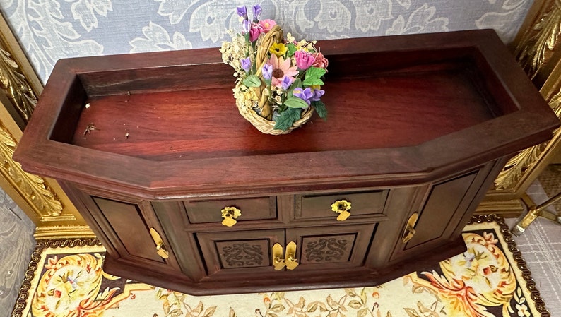 May include: A dark wood cabinet with two doors and two drawers. The cabinet has a carved design on the front and gold-colored handles. A basket of flowers sits on top of the cabinet.