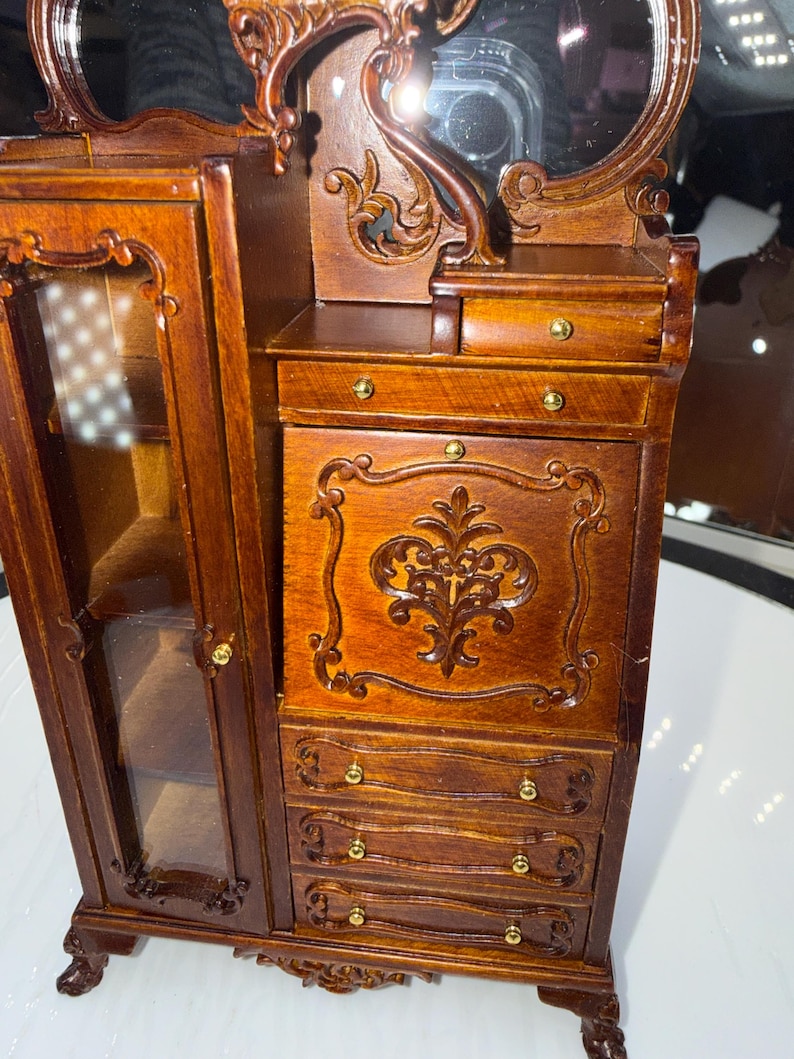 May include: A miniature wooden armoire with a mirror, drawers, and a glass-door cabinet. The armoire is a rich brown color with ornate carvings and gold-colored hardware. The mirror is oval-shaped and sits atop the armoire.