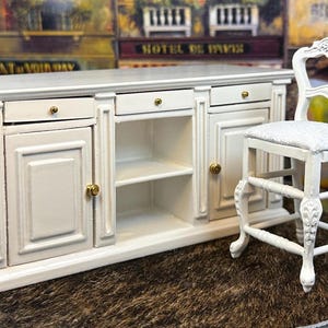 May include: White miniature kitchen island with two doors, two drawers, and open shelves. A white miniature chair with a gray cushion is next to the island.