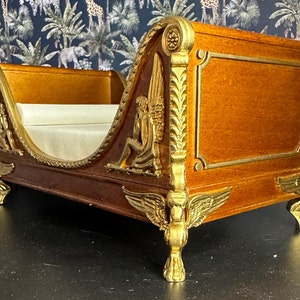 May include: A miniature wooden bed with gold accents and ornate carvings. The bed has a white fabric mattress and is decorated with winged figures.