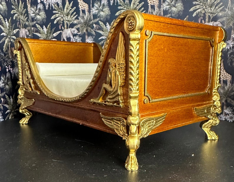 May include: A miniature wooden bed with gold accents and ornate carvings. The bed has a white fabric mattress and is decorated with winged figures.