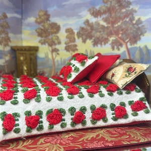 May include: Miniature bed with a white blanket embroidered with red roses and green leaves. The bed has a wooden frame and is adorned with red and floral pillows. The scene is set against a backdrop of a painted landscape.