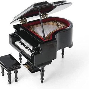 Puede incluir: Un piano de cola negro en miniatura con un banco a juego. La tapa del piano está abierta, revelando un interior rojo y detalles dorados. Las teclas son blancas y negras, y una partitura descansa sobre el atril. Instrumento musical decorativo.