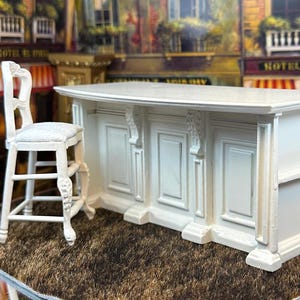 May include: White miniature kitchen island with a curved countertop and three cabinet doors. The island is accompanied by a white miniature bar stool with a carved design.