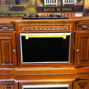 Op de afbeelding: Een miniatuur houten kast met een zwart fornuis en oven. De kast heeft een donkerhouten afwerking met gouden accenten en decoratieve details. De ovendeur staat open en onthult een zwart interieur.