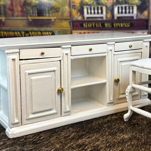 May include: White miniature kitchen island with two doors, two drawers, and a shelf. The island has gold-colored hardware. A white miniature stool with a white cushion is next to the island.