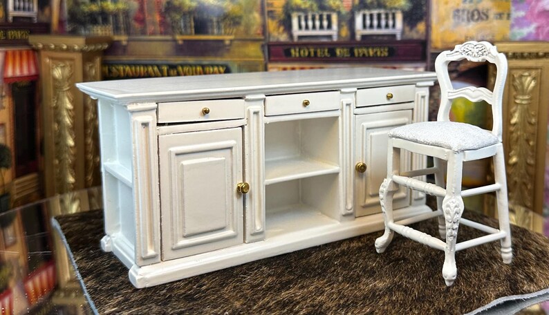 May include: White miniature kitchen island with two doors, two drawers, and a shelf. The island has gold-colored hardware. A white miniature stool with a white cushion is next to the island.