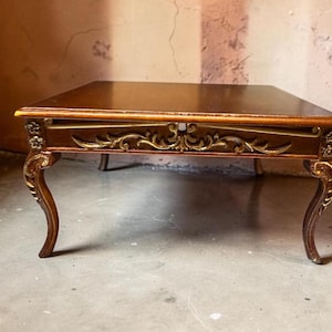 May include: A square coffee table with a dark brown wooden top and a gold-toned apron featuring ornate carved scrollwork and floral details. The table stands on four curved cabriole legs that also feature gold-toned carved accents.
