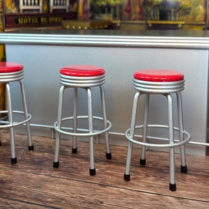 Könnte beinhalten: Drei silberne Barhocker mit roten gepolsterten Sitzen. Die Hocker sind in einer Reihe vor einer silbernen Theke angeordnet.